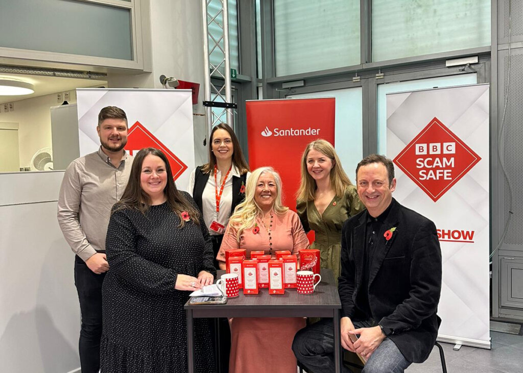 The Santander team attend the BBC Scam Safe pilot in Liverpool. There are four women sitting around a table with a man sat at each end. Behind them is a red advertising board for Santander as well as a board showing the BBC Scam Safe logo which is a red diamond on a white background.