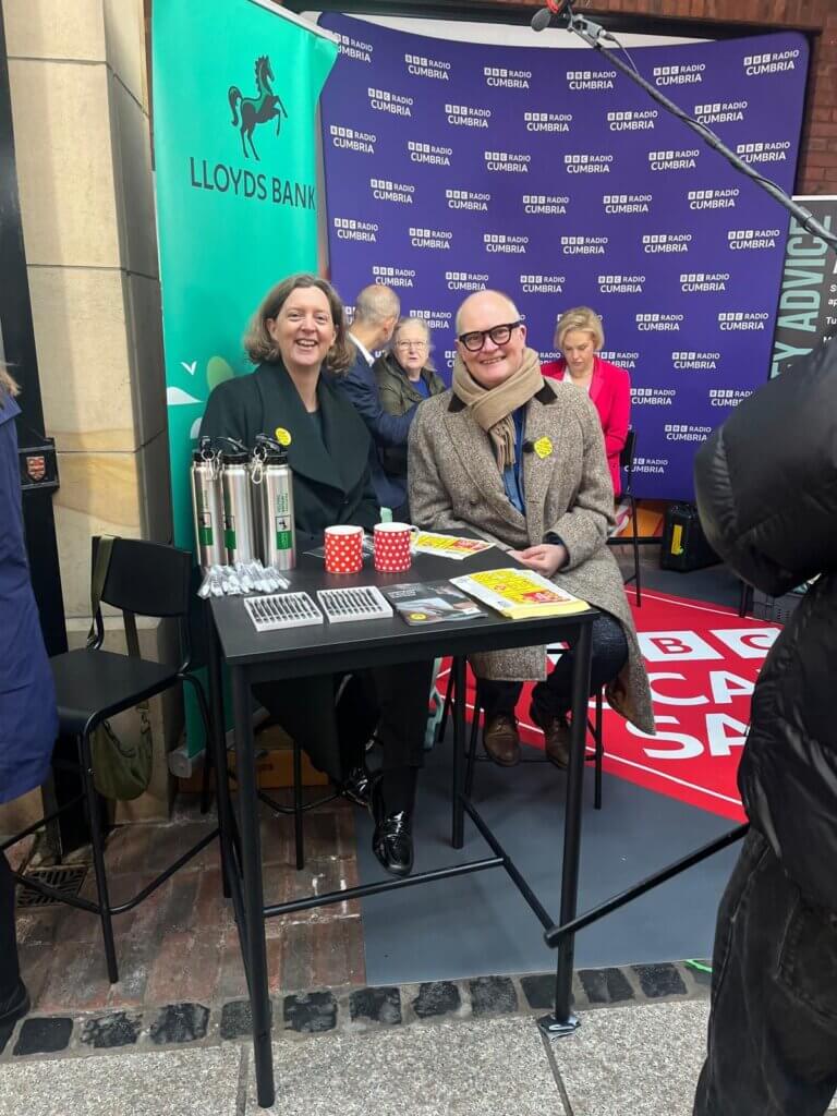 Liz Ziegler sits next to a BBC presenter in front of a BBC Cumbria purple board.