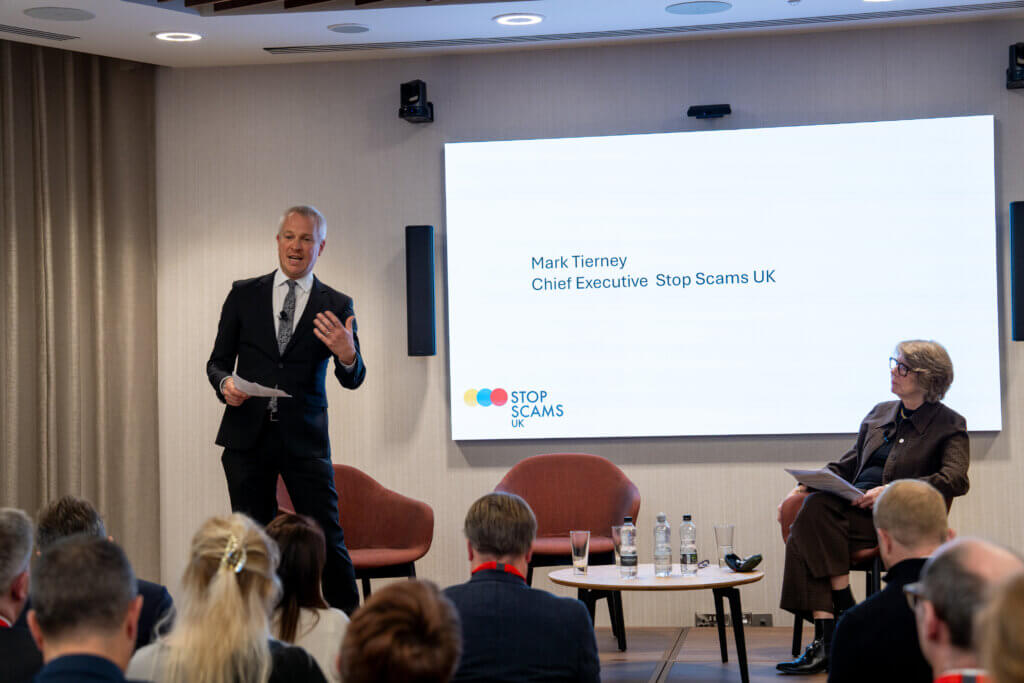 Stop Scams UK CEO Mark Tierney addresses members while standing in front of a white screen with his name and title in the background. Ruth Evans, Chair of SSUK is sat on a chair to his left.