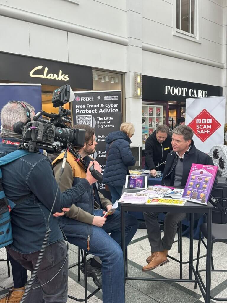 A camera man holds his camera to film Nick Stapleton interview Stuart Skinner of NatWest at a table in a shopping centre.